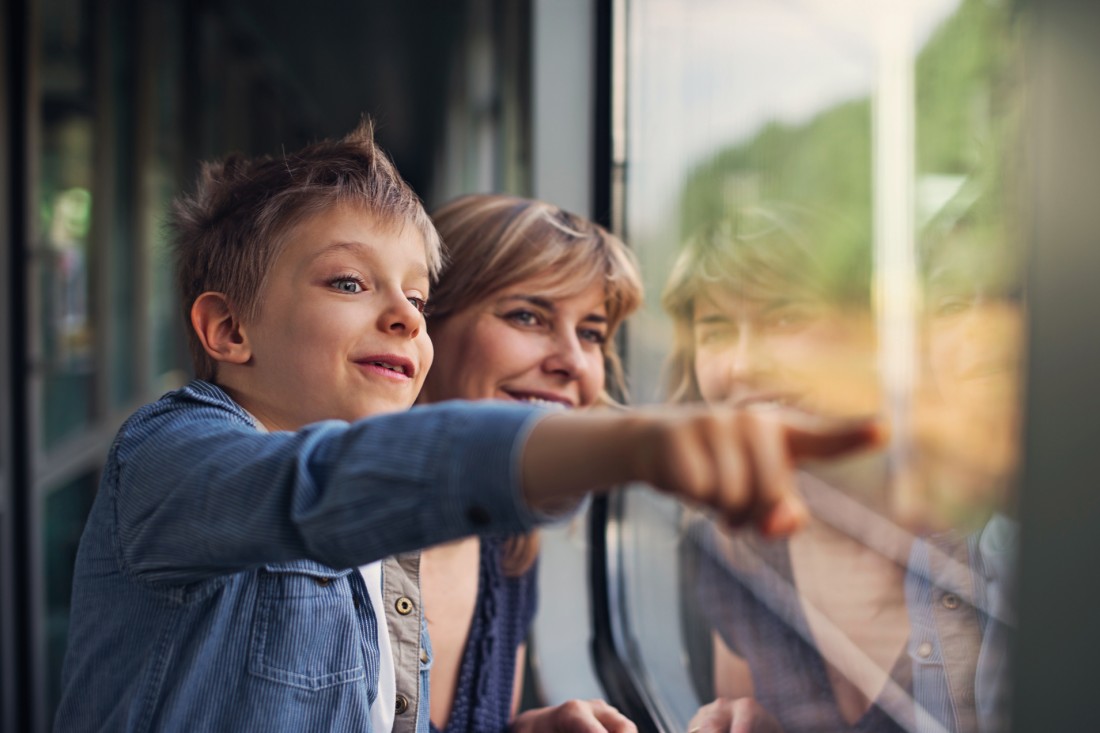 Ein etwa 10-jähriger Junge schaut mit seiner Mutter aus dem Fenster eines Zuges. Er zeigt auf etwas mit ausgestrecktem Arm und Zeigefinger. Er trägt eine blaue Jeansjacke.
