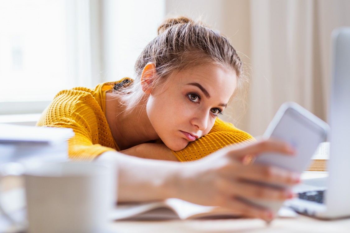 Eine etwa 23-Jährige sitzt am Tisch und schaut lustlos auf ihr Handy.