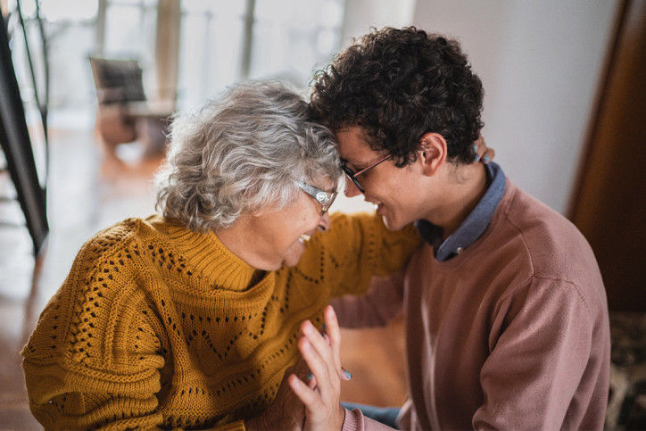 Eine etwa 70-jährige Frau und ihr etwa 20-jähriger Enkelsohn legen innig ihre Köpfe an der Stirn aneinander. Ihre Hand hält ihn sanft im Nacken. Beide lächeln zart. Sie trägt einen senfgelben Strickpullover und Brille, er ein blaues Hemd unter einem blassen roten Sweatshirt und Brille. Sie stehen in einem hellen Wohnraum. 