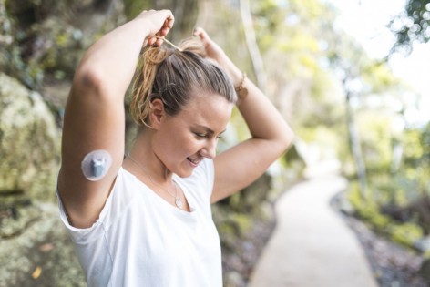 Eine junge Frau mit eine Blutzuckermessgerät am Arm, steht in der Natur und bindet sich ihren Zopf.