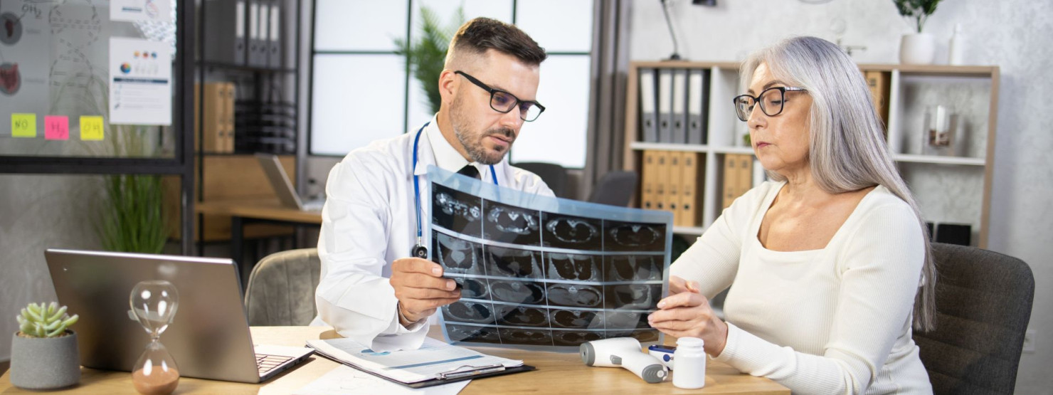Ein Arzt zeigt einer Patientin ihre Röntgenbilder. Beide sitzen am Tisch in der Arztpraxis.