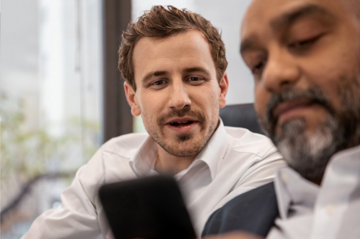 Zwei etwa 35 jährige Männer in weißen Hemden sitzen in einem Büro. Sie schauen zusammen auf ein Smartphone.