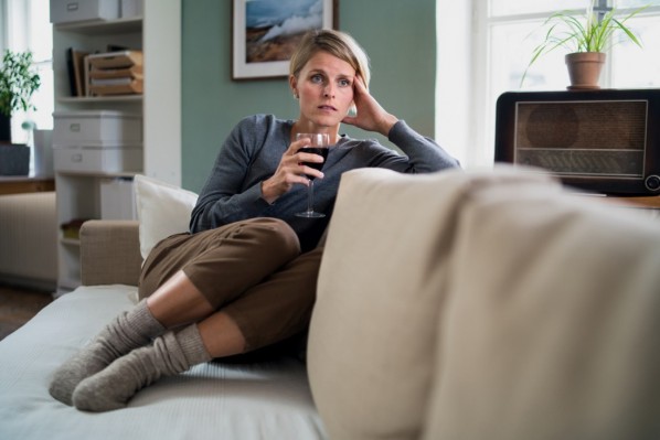 Frau Mitte 40 sitzt auf dem Sofa und hat ein Glas Rotwein in der Hand. Sie schaut besorgt.