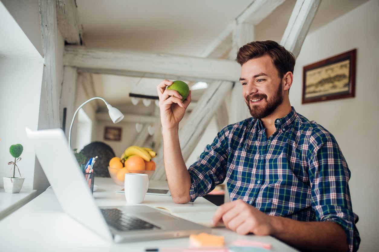 Eine etwa 40 jähriger Mann sitzt am Laptop im Homeoffice und hält einen Apfel in der Hand