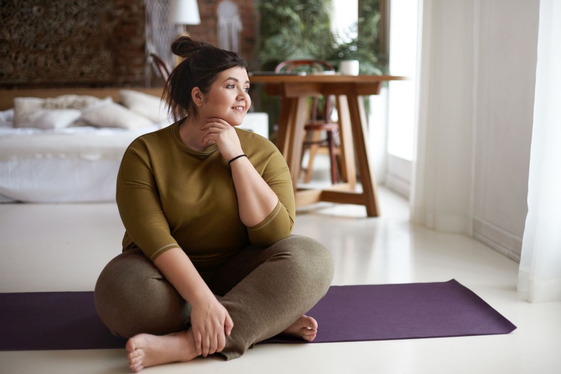 Eine mollige Frau sitzt auf ihrer Yogamatte und schaut interessiert aus dem Fenster.