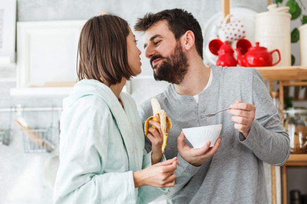 Ein etwa 30-jähriges Paar steht zusammen in der Küche. Er hält eine Schüssel Müsli, sie eine Banane.