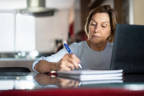 Eine etwa 55-jährige Frau sitzt am Tisch und macht sich Notizen. Vor ihr steht ein Laptop.