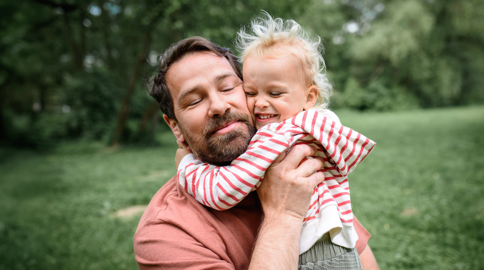 Ein Mann, etwa Mitte 30, steht mit seinem Kleinkind im Arm in einer grünen Umgebung mit Bäumen im Hintergrund. Er hält das Kind unter den Armen, das Kind hat die Arme um den Hals Hals des Vaters gelegt. Sie drücken ihre Wangen liebevoll aneinander. Beide haben die Augen geschlossen und lächeln.