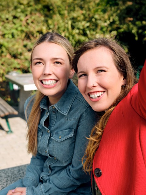 Die Auszubildenden Alina und Josi sitzen in einem Park auf einer Tischtennisplatt und machen gemeinsam ein Selfie.