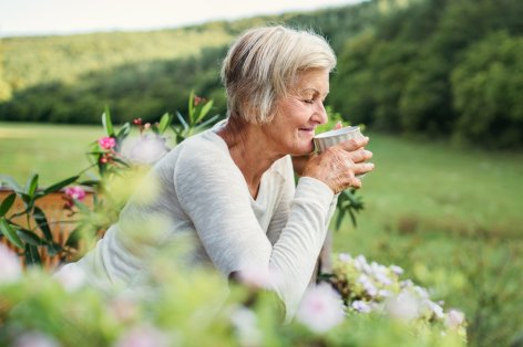 Eine Seniorin steht an einen Zaun gelehnt und trinkt eine Tasse Tee. Sie schaut in die Natur und lächelt.