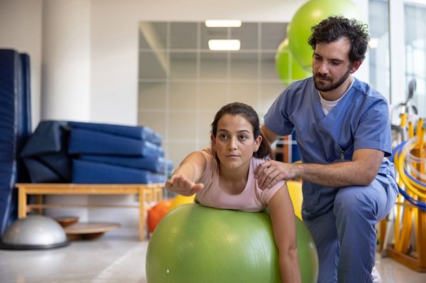 Physiotherapist helping a woman in physical therapy doing stretching exercises using a fitness ball