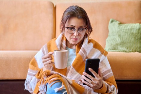 Eine Frau sitzt vor einem Sofa und schaut auf ihr Handy. Sie ist krank und hält ein Tasse in ihrer Hand.
