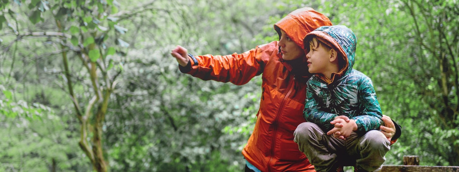 Eine etwa 40-jährige Mutter zeigt ihrem etwa 6-jährigen Sohn etwas mit Regenkleidung im Wald.