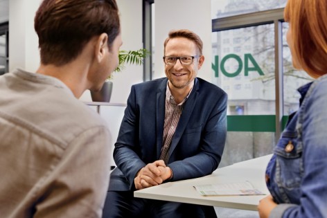 Ein etwa 40 jähriger Mann mit Brille und blauem Jakett unterhält sich mit 2 Personen. Sie befinden sich in einer AOK Filiale. An der Scheibe im Hintergrund ist das AOK-Logo angebracht.