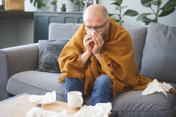 Ein etwa 40 jähriger Mann sitzt auf dem Sofa und schnäuzt in ein Taschentuch. Er ist erkältet.