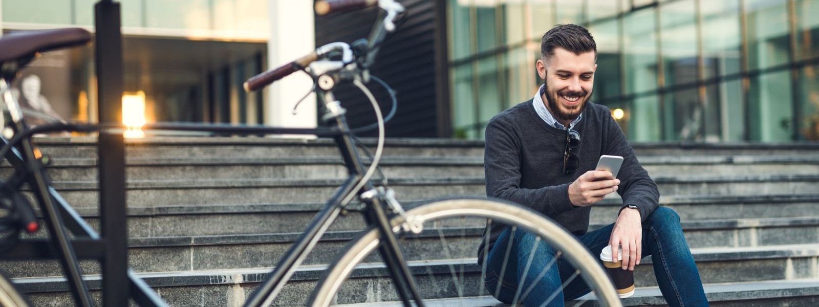 Ein etwas 30 jähriger Mann sitzt auf einer Treppe vor einem Bürogebäude. Er schaut auf sein Handy und lächelt. Vor ihm steht ein Fahrrad.