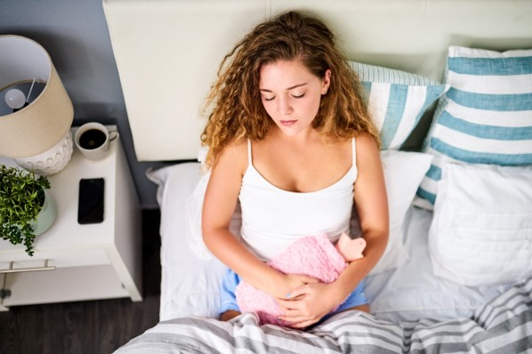 Eine dunkelblonde Frau mit Locken hält sich eine Wärmflasche auf den Bauch, während sie im Bett liegt.