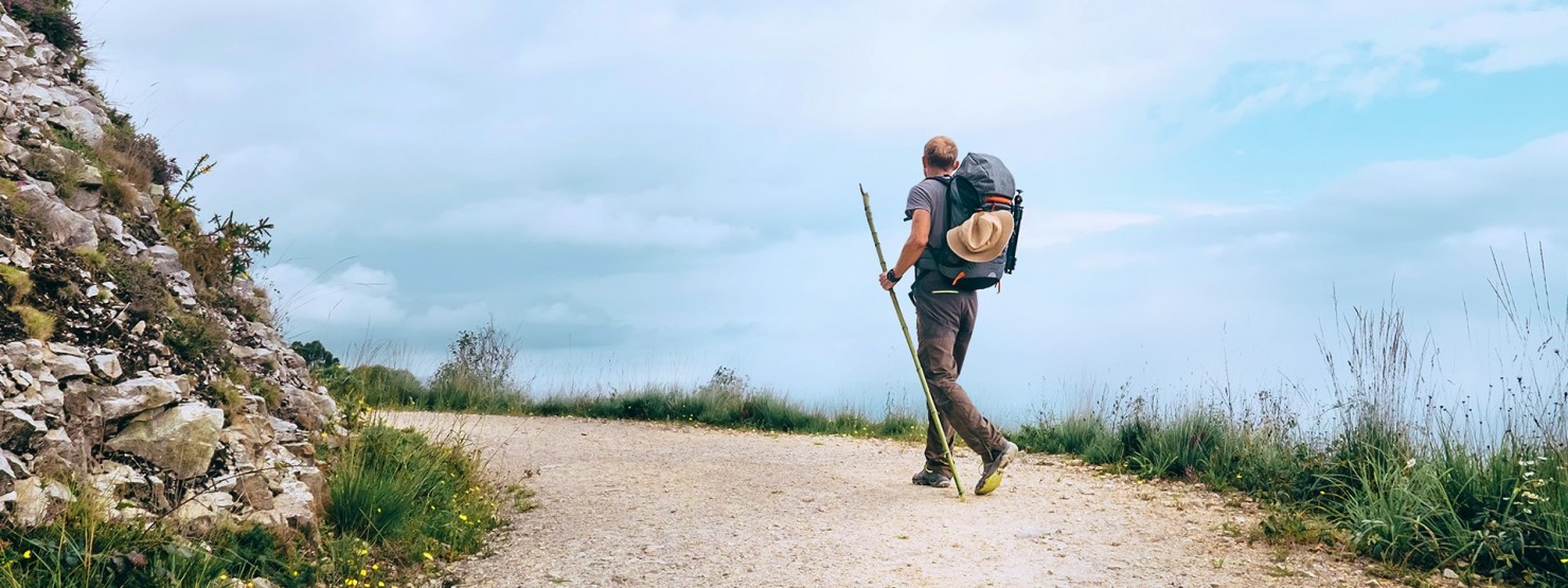 Mann mit Rucksack und Wanderstock läuft auf einem Weg in Richtung Horizont