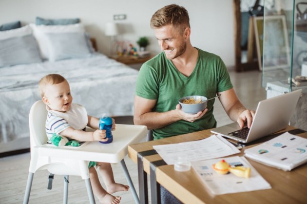 Father using laptop to finish some work from home and baby drinking from baby bottle, they are at kitchen table