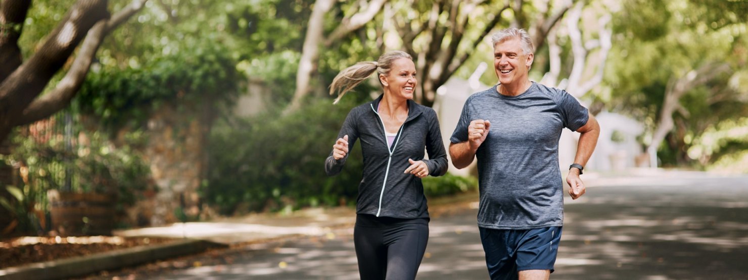 Ein Mann und eine Frau, beide etwa 50, joggen gemeinsam auf einer von Bäumen umsäumten Straße.