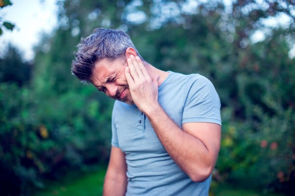 Ein etwa 35 jähriger Mann steht in einem grünen Garten und fasst sich mit schmerzverzerrtem Gesicht an sein linkes Ohr. 
