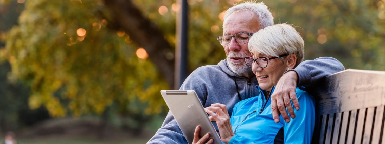 Ein Seniorenpärchen sitzt in der Natur gemeinsam auf einer Holzbank. Der Mann umarmt die Frau. Beide schauen gemeinsam auf ein Tablet, welches die Frau in der Hand hält.
