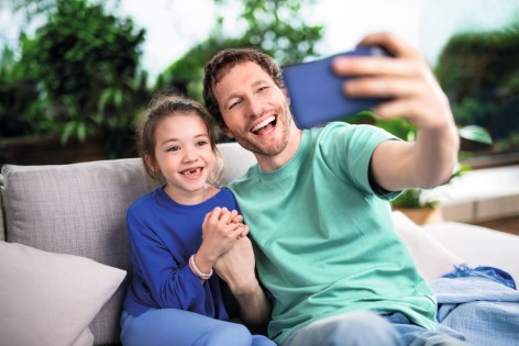 Vater und Tochter sitzen auf der Couch und lächeln breit in die Handykamera.