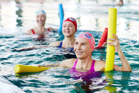 Drei Frauen befinden sich in einer Schwimmhalle im Wasser und machen Wassergymnastik mit bunten Badenudeln.
