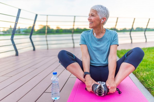 Eine etwa 60 jährige Frau sitzt im Schneidersitz auf einer Sportmatte am Rande eines Sees. Neben ihr steht eine kleine Wasserflasche. Sie schaut lächelnd aufs Wasser und dehnt dabei ihre Beine.