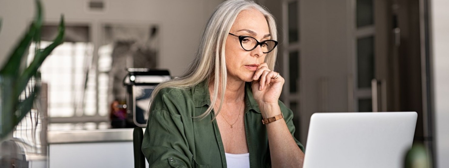 Eine etwa 60 jährige Frau sitzt an einem Tisch und arbeitet an einem Laptop. Neben ihr liegen ihre Notizen.