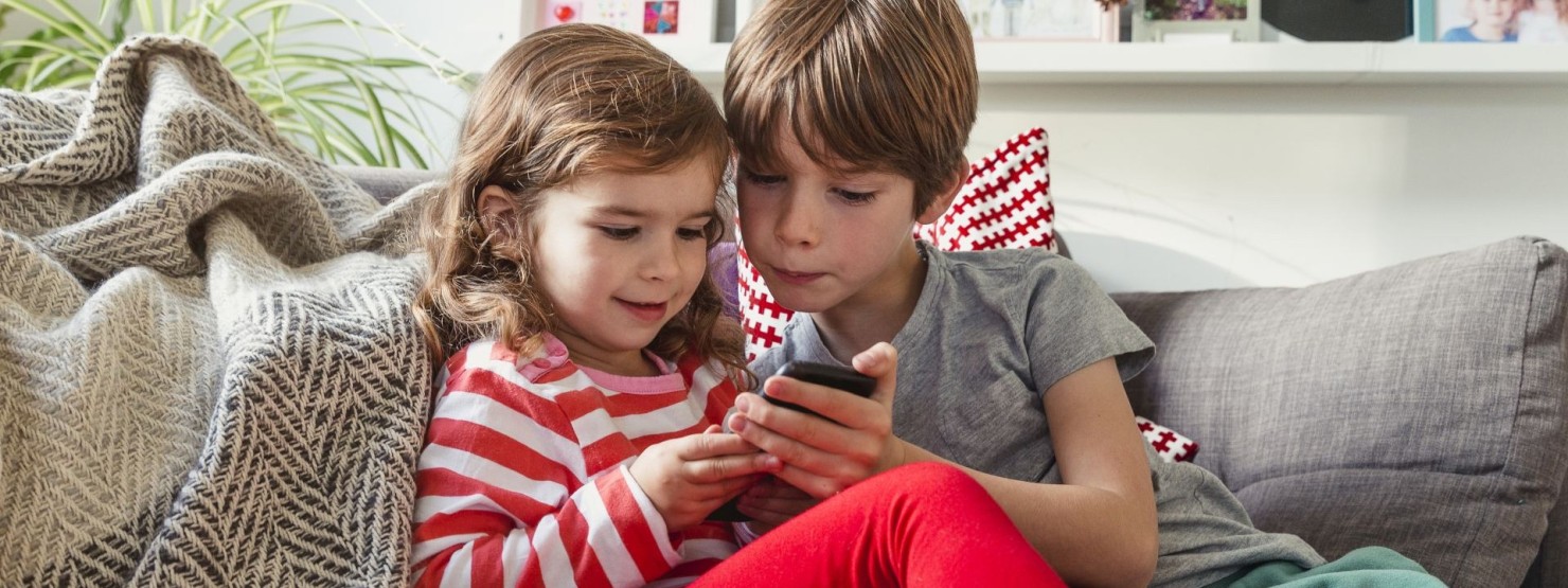 Zwei Kinder schauen gemeinsam auf ein Handy und sitzen auf einem Sofa.