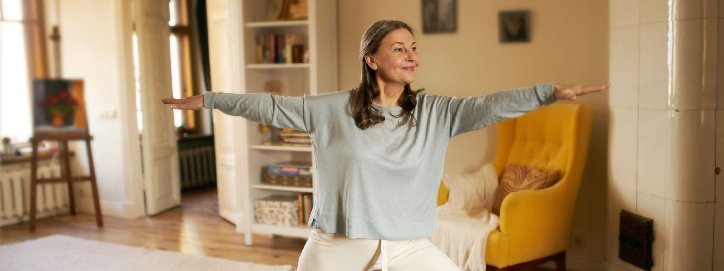 Eine etwa 60jährige Frau steht in einem Wohnzimmer auf einer Yogamatte und macht eine Übung mit ausgestreckten Armen.